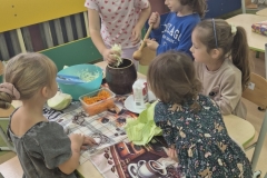 3. Przy stoliku przedszkolnym siedzi troje przedszkolaków a dwoje stoi. Dziewczynka w białej bluzce z serduszkami wkłada do glinianej beczki garść poszatkowanej kapusty. Stojący obok chłopiec trzyma drewnianą pałkę do ubijania. Pozostałe dzieci przyglądają się tym działaniom. Na stoliku na kolorowych serwetach widać pojemnik z poszatkowaną kapustą drugi ze startą marchwią i woreczek z solą. W tle okna przedszkolne ozdobione kolorowymi liśćmi i sylwetami jeża.