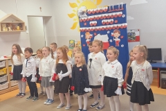 Sala przedszkolna . Grupa dzieci ubrana w strojach galowych stoją przed tablicą okolicznościową na której znajduje się głowa orła , napis w sercach biało- czerwonych PEŁNI WDZIĘCZNOŚCI ZA DAR NIEPODLEGŁOŚCI , postacie chłopca i dziewczynki z biało – czerwonymi chorągiewkami . Pomiędzy postaciami kontur mapy polski i kwiatki biało- czerwone. Dzieci śpiewają piosenkę.