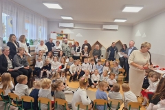 Sala przedszkolna . Dorośli i dzieci . Dzieci siedzą na krzesełkach . Dorośli stoją ; tylko dwoje z nich siedzi na krzesłach. Część dorosłych trzyma białe prostokątne kartki . Dorośli śpiewaja pieśni patriotyczne , a dzieci słuchają.