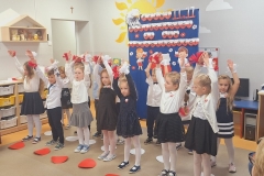 .Sala przedszkolna . Grupa dzieci ubrana w strojach galowych stoją przed tablicą okolicznościową na której znajduje się głowa orła , napis w sercach biało- czerwonych PEŁNI WDZIĘCZNOŚCI ZA DAR NIEPODLEGŁOŚCI , postacie chłopca i dziewczynki z biało – czerwonymi chorągiewkami . Pomiędzy postaciami kontur mapy polski i kwiatki biało- czerwone. Dzieci z uniesionymi rękami do góry.Na rękach mają białe i czerwone pompony zrobione z bibuły . Przed dziećmi na podłodze leżą białe i czerwone koła.