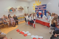 Sala przedszkolna . Grupa dzieci ubrana w strojach galowych stoją przed tablicą okolicznościową na której znajduje się głowa orła , napis w sercach biało- czerwonych PEŁNI WDZIĘCZNOŚCI ZA DAR NIEPODLEGŁOŚCI , postacie chłopca i dziewczynki z biało – czerwonymi chorągiewkami . Pomiędzy postaciami kontur mapy polski i kwiatki biało- czerwone. Z prawej strony stoi pani w okularach , w cienmych spodniach i pstrokatej bluzce - przemawia mikrofonu. Z lewej strony widać dwie panie opatre o pólki indywidualne dzieci w strojach odświętnych.