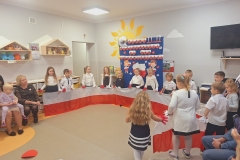 Sala przedszkolna . Grupa dzieci ubrana w strojach galowych stoją w półkolu trzymając w dłoniach długi biało- czerwony materiał przypominający flagę Polski. Z lewej strony siedzi na krzesełku pani ubrana w ciemną sukienkę.