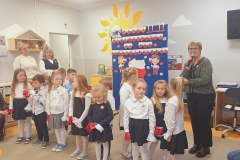 Sala przedszkolna . Grupa dzieci ubrana w strojach poruszają się wężykiem trzymając w dłoniach długi biało- czerwony materiał przypominający flagę Polski.Z lewej stronie trzy panie siedzą na niskich przedszkolnych krzesełkach ; dwie z nich trzymają na kolanach małe dzieci. W tle widać półki indywidualne dla dzieci , a na ścianie dwie wiszące szafki z pluszakami.