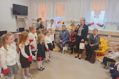  Sala przedszkolna . Z lewej strony widać część dzieci ubranych na galowo.Z prawej strony mężczyzna w niebieskim garniturze siedzi na krześle . Obok niego z prawej strony widać trzy kobiety ; jedna z nich s toi i trzyma w jednej ręce mikrofon a w drugiej papierową kolorową torebkę na prezenty . Pozostałe panie siedzą na krzesłach. Z prawejstrony mężczyzny za dziecmi stoją dwie panie. W tle widać okna przysłonięte firankami i roletami . Na parapetach okien kwiaty i biało- czerwone chorągiewki . Na ścianie widać czarny ekran telewizora.