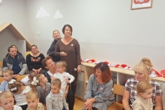 Sala przedszkolna . Dzieci w strojach odświętnych siedzą na krzesełkach. Za dziećmi sześć dorosłych osób . Dwie z nich stoją , jedna ubrana na brązowo trzyna w ręce mikrofon .Z prawej strony dwie panie siedzą na niskich krzesełkach ; jedna z nich trzyma na kolanach dziecko . W tle szara ściana , na ścianie widać fragment godła Polski.