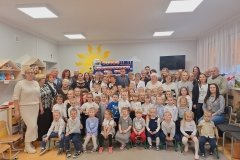 Sala przedszkolna . Dorośli i dzieci ustawieni w zwartej grupie pod ścianą , na której widać część napisu w sercach biało- czerwonych PEŁNI WDZIĘCZNOŚCI ZA DAR NIEPODLEGŁOŚCI.Dzieci ustawieni przed dorosłymi . Część z grupy dzieci siedzi na krzesełkach . Na twarzach wszystkich zebranych maluje się uśmiech i radość.