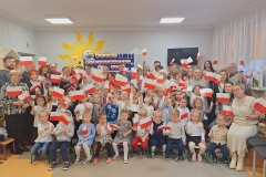 Sala przedszkolna . Dorośli i dzieci ustawieni w zwartej grupie pod ścianą , na której widać część napisu w sercach biało- czerwonych PEŁNI WDZIĘCZNOŚCI ZA DAR NIEPODLEGŁOŚCI. Za tablicą widać część żółtego dużego słonka . Z prawej strony na ścianie widać czarny ekran telewizora . Dzieci ustawiene przed dorosłymi . Część z grupy dzieci siedzi na krzesełkach . Na twarzach wszystkich zebranych maluje się uśmiech i radość. Każda z osób trzyma biało- czerwoną chorągiewkę.