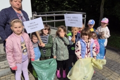 3. Grupa dzieci przedszkolnych ubrana w kolorowe kurtki i spodnie stoi na kostce brukowej przed budynkiem przedszkola. Za nimi widoczna jest balustrada. Z lewej strony stoi nauczycielka w ciemnym swetrze, za nią widoczny jest fragment jasnej ściany budynku a z prawej zielone drzewa i fragment nieba. Dwie dziewczynki po lewej stronie trzymają zielony worek, za nimi stoi chłopiec          z transparentem: Akcja Sprzątania Świata. Z prawej strony dziewczynka trzyma worek żółty, za nią chłopiec z transparentem: Małe dzieci segregują śmieci. Transparenty są białe, na nich napisy drukowanymi czarnymi literami.