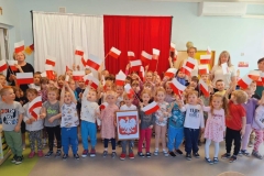 Grupa dzieci stoi  w trzech rzędach na tle biało- czerwonej dekoracji. Każde trzyma w rękach chorągiewki.