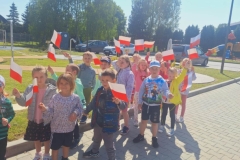 Grupa dzieci w kolorowych strojach stoi na szarym chodniku. Każde dziecko trzyma w ręce biało- czerwoną chorągiewkę.  W tle szaro- żółta altanka.