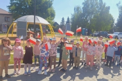 Grupa  dzieci stoi na chodniku. Każde z nich trzyma w ręku biało- czerwoną chorągiewkę.