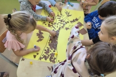 2. W sali przedszkolnej przy stoliku siedzi grupa dzieci. Wyklejają one brązowymi kawałkami papieru sylwetę drzewa narysowaną na żółtym kartonie. W tle widoczne kolorowe, drewniane obudowy kaloryferów.