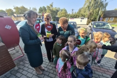 4. Na chodniku stoją trzy kobiety. Dwie z nich trzymają w ręku ozdobne serduszka - kwiaty w kolorowych pojemnikach. Obok stoi grupa dzieci przedszkolnych. Za nimi widoczne są zaparkowane samochody, dom, paczkomat, budynek sklepu. W tle widac drzewa.