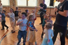 Dzieci ubrane  w kolorowe  stroje stoją w rozsypce na parkiecie sali gimnastycznej.  Widać też dwóch panów ubranych w czarne koszulki i spodnie. W tle widać duże  okna.