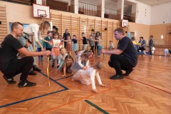 Dwóch panów kuca na parkiecie. W rękach  trzymają złoty sznurek. Pod sznurkiem przechodzą dzieci.