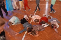 Siedmioro dzieci lezy na podłodze. Pozostałe trzymają się za ręce tworząc półkole.