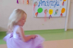 Dziewczynka we fioletowej sukience tańczy w koło, w tle kolorowa tablica.