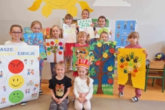 Grupa 9 dziewczynek stoi na podłodze. W rękach trzymają plakaty przedstawiające drzewka emocji. Przed nimi klęczą 2 dziewczynki. Jedna z nich ma na głowie koronę.