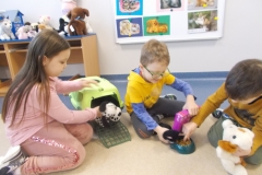 Troje dzieci bawi się pluszowymi zwierzętami. Dziewczynka w różowym stroju  ,,wyprowadza’’ psa z transportera w kierunku chłopca ubranego w żółto - niebieską bluzę i szare spodnie. Sypie on suchą karmę do miski. Klęczący obok chłopiec  w ciemno żółtej bluzie bierze w palce kawałek karmy, w drugiej ręce trzyma białego pluszowego psa. W tle widać tablicę z planszami psów i kotów oraz półkę z ustawionymi pluszowymi zwierzętami.