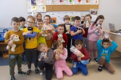W sali przedszkolnej znajduje się ustawiona na tle ściany grupa kilkunastu dzieci. Czworo z nich klęczy na przodzie, pozostałe stoją za nimi. Wszystkie dzieci trzymają w rękach pluszowe koty lub psy, tulą je do siebie. Po prawej stronie stoi biurko, na nim laptop. W tle tablica z literami.