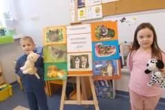 Do stelaża przypięta jest korkowa tablica, na której znajdują się dwa napisy drukowanymi czarnymi literami na białym papierze: ,, Pomagamy’’ i pod spodem ,, Wspieramy Tarnowski Azyl’’. Naokoło napisów przypięte są kolorowe plansze z wizerunkami kotów i psów. Z prawej strony stoi dziewczynka w różowym stroju, w ręce trzyma pluszowego dalmatyńczyka. Z lewej strony stoi chłopiec ubrany na niebiesko. W ręce trzyma białego kota. W tle tablica z literami.