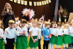 Dzieci stoją w półkolu.  Dziewczynki ubrane w białe bluzeczki i niebieskie spódniczki. Chłopcy w białe koszule i ciemne spodnie. Za nimi na ciemnym tle napis Dzień Babci i Dziadka.