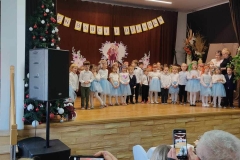 Dzieci stoją  w szeregu. Dziewczynki ubrane w białe  bluzeczki I zielone spódniczki,  chłopcy w białe  koszule i ciemne spodnie. Za nimi pani w ciemnej sukience.