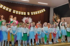 Dzieci stoją w półkolu.  Dziewczynki ubrane w białe bluzeczki i niebieskie spódniczki. Chłopcy w białe koszule i ciemne spodnie. Za nimi na ciemnym tle napis Dzień Babci i Dziadka.