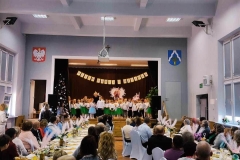 Na sali ustawione dwa rzędy stołów przy nich na krzesłach siedzą  panie i panowie. W dali na scenie widać grupę dzieci, a za nimi napis Dzień Babci i Dziadka.