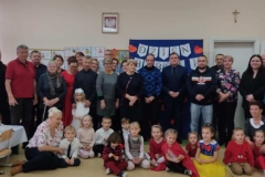 Babcie, dziadkowie , zaproszeni goście stoją w dwóch rzędach, przed nimi siedzą na podłodze dzieci