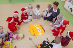 Dzieci siedzą na podłodze wokół garnka, uderzają łyżkami drewnianymi o podłogę w rytm muzyki.