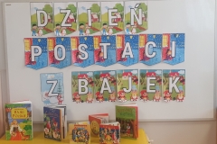 Sala przedszkolna. Na białej prostokątnej tablicy kolorowy napis DZIEŃ POSTACI Z BAJEK. Pod tablicą ustawione książki z bajkami dla dzieci.