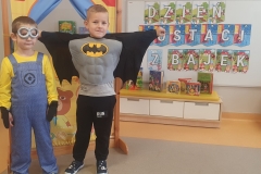 Sala przedszkolna.Dwójka chłopców w strojach ; Jeden przebrany za Batmana, drugi za Minionka stoją przed teatrzykiem, za mimi tablica z napisem DZIEŃ POSTACI Z BAJEK. Chłopiec przebrany za Batmana ma rozłożone ręce w bok.
