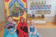 Sala przedszkolna. Czwórka dzieci w strojach ; Smoka , księżniczki , Elsy i biedronki. Dziewczynki siedzą na podłodze przed teatrzykiem , w okienku teatrzyku stoi chłopiec przebrany za smoka , za mimi tablica z napisem DZIEŃ POSTACI Z BAJEK.