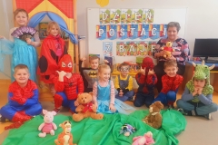 Sala przedszkolna. Przed tablicą dzieci w strojach postaci bajkowych . Dwie dziewczynki stoją , pozostałe dzieci siedzą na podłodze przed tablicą . Przed dziećmi leży zielony materiał , na nim postacie z bajki Kubuś Puchatek . Z lewej strony za stojącymi dziewczynkami widać przedszkolny teatrzyk. Z prawej strony tablicy na krzesełku siedzi wychowawczyni , na kolanach trzyma pluszaka – Kubusia Puchatka.. W tle tablica z napisem DZIEŃ POSTACI Z BAJEK.