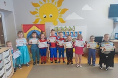 Sala przedszkolna. Przed tablicą stoją dzieci w strojach postaci bajkowych z pamiątkowymi dyplomami.