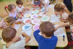 Przy stolikach siedzą dzieci, na stole kolorowa sól na talerzykach, dzieci nakładają sól na ilustracje motyla i balona.