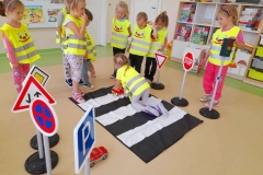 Grupa dzieci ubrana w kamizelki odblaskowe, stoi wokół biało czarnych pasów, dziewczynka przejeżdża przez pasy samochodem zabawkowym. Wokół pasów ustawione znaki i sygnalizacja świetlna.