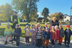 Dzieci stoją w dwuszeregu.  Za nimi pani ubrana w ciemny strój.  Obok stoi pan policjant.  W tle zielona trawa i drzewa.