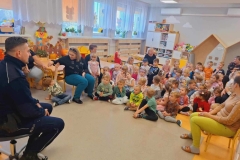 Duża grupa dzieci siedzi na podłodze. Przed  nimi na krześle siedzi policjant ubrany w granatowy strój.  W tle okna i jasna ściana.