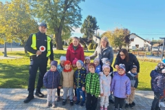 Dzieci stoją w dwuszeregu.  Obok stoi pan policjant.  W tle zielona trawa i drzewa.