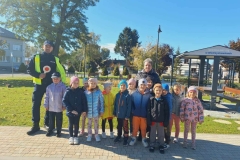 Dzieci stoją w dwuszeregu.  Za nimi pani ubrana w ciemny strój.  Obok stoi pan policjant.  W tle zielona trawa i drzewa.