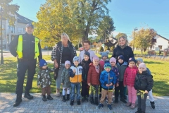 Dzieci stoją w dwuszeregu.  Za nimi stoją trzy panie,  a obok pan policjant. W tle zielona trawa i drzewa.