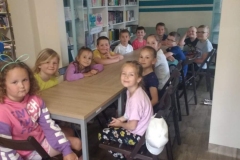 Dzieci siedzą w czytelni przy stoliku. Za nimi przy ścianie stoi szafa z półkami, na których poukładane są książki.