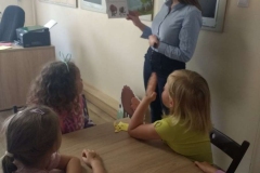 Pani w niebieskiej bluzce i ciemnych spodniach stoi przy ścianie. Pokazuje obrazki w książce. Dzieci siedzą przy stoliku i patrzą w jej stronę.