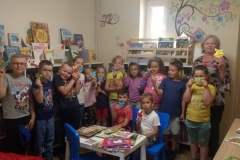 Grupa dzieci i pani stoją w półkolu. Przed nimi przy stoliku na niebieskich krzesełkach siedzi 2 chłopców. Wszyscy trzymają w rękach kolorowe zakładki do książki. Na ścianie namalowana gałązka z kwiatami, a wśród nich sowa.