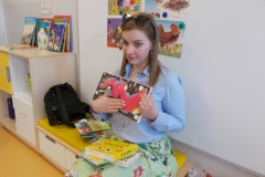 W sali przedszkolnej na szafce - siedzisku z żółtym materacem siedzi pani z biblioteki. Ubrana jest w błękitną koszulową bluzkę i zieloną spódnicę w kwiaty. W ręku trzyma otwartą książkę i prezentuje dzieciom sensoryczny obrazek. Na jej kolanach leżą trzy książki, ta na wierzchu ma kolor żółty i widoczny tytuł ,, A to kto?’’. Po jej lewej stronie leżą kolejne książki, za nimi czarna torebka. Z lewej strony widoczna jest szafka z wyłożonymi książkami, w tle biała tablica z obrazkami.