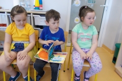 W sali przedszkolnej na krzesełkach siedzi troje dzieci. Dziewczynka z prawej strony ma jasne spodnie w kwiaty i błękitną bluzkę z różowym nadrukiem. Dziewczynka z lewej ubrana jest w żółtą koszulkę i niebiesko - żółtą spódniczkę. Chłopiec siedzący w środku ma ciemne spodnie i niebieską koszulkę. Przegląda książkę. W tle widoczne drzwi wejściowe na salę i szafki przedszkolne.