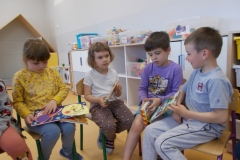 Czworo dzieci siedzi na krzesełkach w sali przedszkolnej. Ubrane są w kolorowe stroje. Każde z nich trzyma książkę. Dziewczynka ją przegląda. Chłopiec siedzący po prawej stronie pokazuje swoją książkę pozostałym. W tle widoczne szafki przedszkolne z zabawkami.
