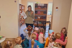 Grupa dzieci siedzi na kolorowych pufach. Przed nimi stoi pani w beżowym żakiecie. W prawej ręce trzyma kartkę z tekstem i obrazkami. Za nią półka z książkami i brązowe krzesło. Na ścianie kolorowe literki.